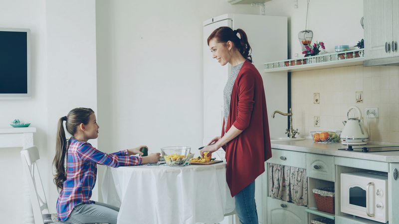 Mãe prepara o lanche enquanto a filha conversa com ela à mesa da cozinha